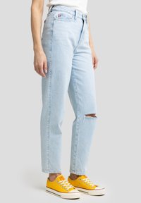 Jeans de tiro alto en azul claro con un corte relajado y una rodilla desgastada. Combinados con zapatillas de lona amarillas y una blusa ajustada blanca.
