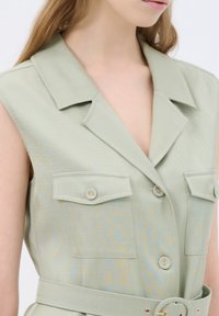 Chemise sans manches vert clair avec un col, comportant deux poches poitrine et une ceinture à la taille. Texture lisse et motif subtil sur l'ensemble.