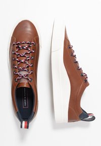 Brązowe skórzane sneakersy z białą gumową podeszwą, kolorowymi plecionymi sznurowadłami i granatowym wnętrzem. Posiadają paskowany akcent na pięcie.