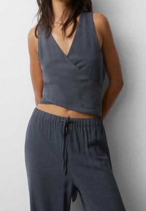 Kobieta ubrana w bezrękawnikowy, ciemnoszary crop top typu wrap i dopasowane spodnie ze sznurkiem, stojąca na jednolitym białym tle.