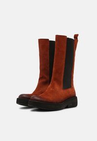 Brązowe zamszowe botki za kostkę z czarnymi elastycznymi panelami po bokach, okrągłym noskiem i niską, płaską czarną podeszwą. Pętla z tyłu ułatwiająca zakładanie.
