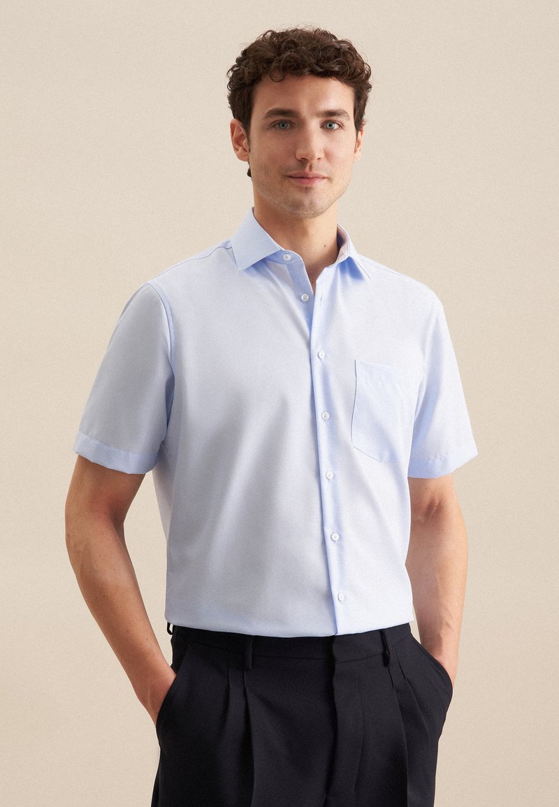 Camicia azzurra a maniche corte in tessuto morbido con colletto a punta, sette bottoni e tasca sul petto. Indossata con pantaloni scuri, fit casual.