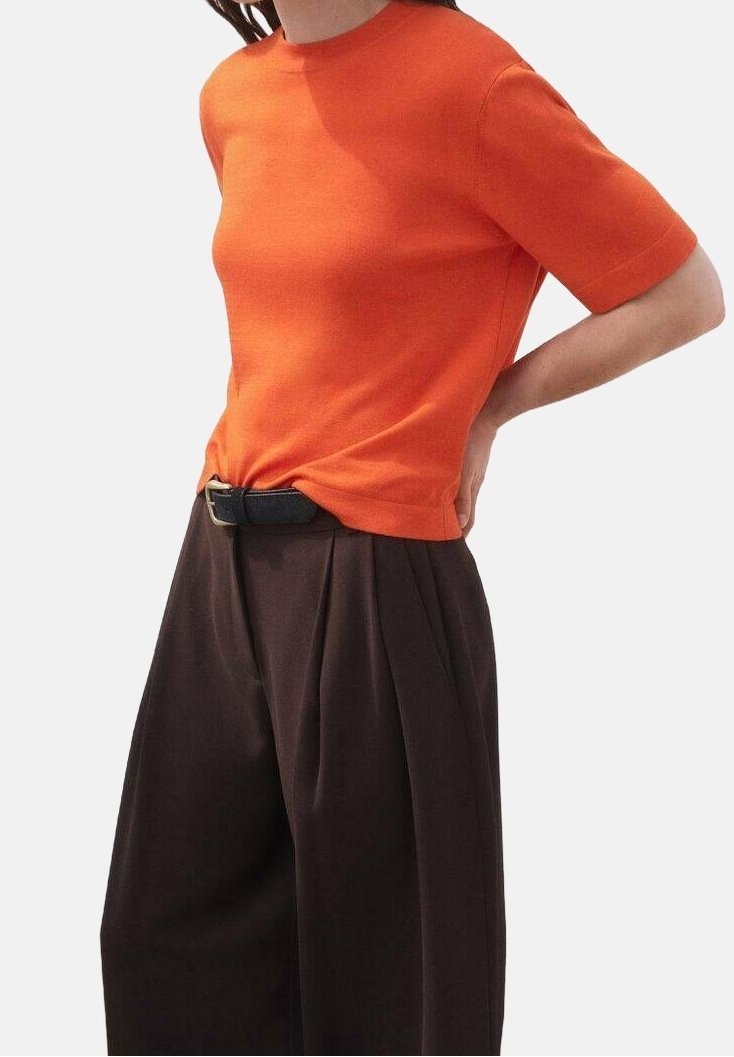 Femme portant un haut orange ajusté à manches courtes rentré dans un pantalon large taille haute marron foncé avec ceinture noire.