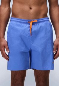 Μπλέ μαγιό με πορτοκαλί κορδόνι που φοράει ένα άτομο που στέκεται με τα χέρια χαλαρά στα πλάγια μπροστά σε ένα μονόχρωμο φόντο.