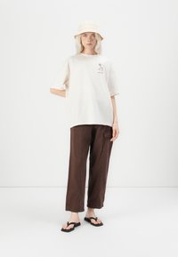 Beige oversized t-shirt with logo, brown wide-leg pants, and a light tan bucket hat. Black flip-flops complete the outfit.