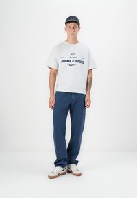 Lichtgrijze katoenen t-shirt met de tekst "ATHLETICS" in marineblauw, gecombineerd met blauwe jeans. Extra witte sneakers en een donkere pet maken de look compleet.