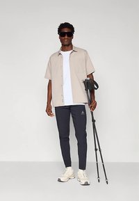 Svetlo bež srajca s kratkimi rokavi, čez belo majico, temno modre hlače in bele superge. Oseba drži črno sprehajalno palico z ročaji.