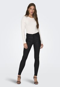 Zwarte skinny jeans met een hoge taille en een gladde textuur, stijlvolle combinatie met een lange mouw witte top. Model draagt zwarte hakken met bandjes.