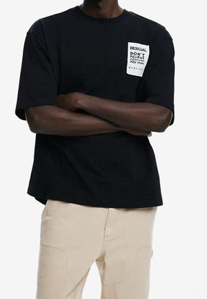 Uomo con le braccia incrociate che indossa una maglietta nera a maniche corte con una toppa bianca su cui è scritto "Desigual Don't Believe Everything You Think" e pantaloni beige.