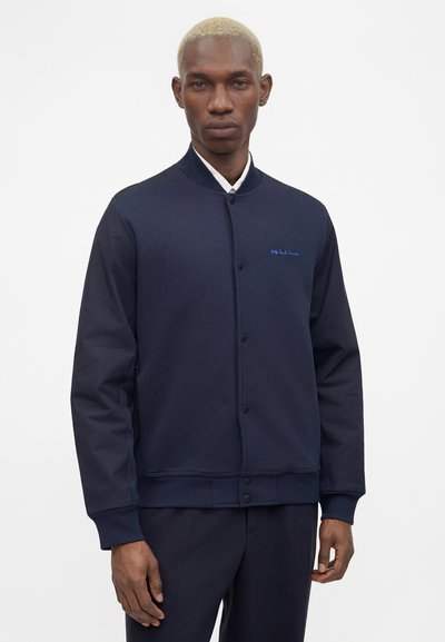 Άνδρας με κοντά ξανθά μαλλιά, φορώντας μπλε σκούρο σακάκι με κουμπιά και αντίστοιχο παντελόνι μπροστά σε απλό λευκό φόντο.