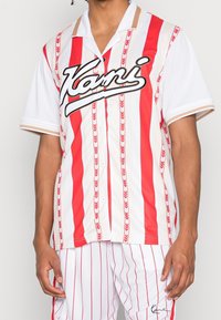 Camisa de mangas cortas a rayas rojo y blanco con botones en el frente, que presenta un logo en negro y patrones de flechas en las rayas.