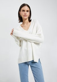 Biały, oversize'owy sweter z dzianiny z dekoltem w serek, ściągaczami przy mankietach i u dołu. Połączony z jasnoniebieskimi dżinsami, prezentowany na neutralnym tle.