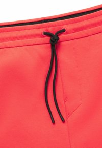 Pantaloni della tuta color corallo con vita regolabile. Presentano lacci neri e una texture di tessuto liscia. Inclusa una tasca laterale.
