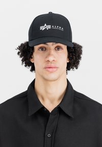 Melnas beisbola cepures ar plakano malu, ar baltu logo "ALPHA INDUSTRIES" priekšā. Valkāta ar melnu apkakles krekls.