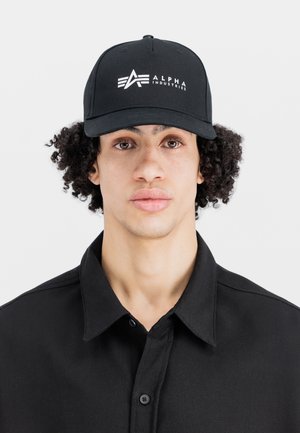 Cappellino da baseball nero con visiera piatta, caratterizzato da un logo bianco "ALPHA INDUSTRIES" sulla parte anteriore. Indossato con una camicia nera con colletto.