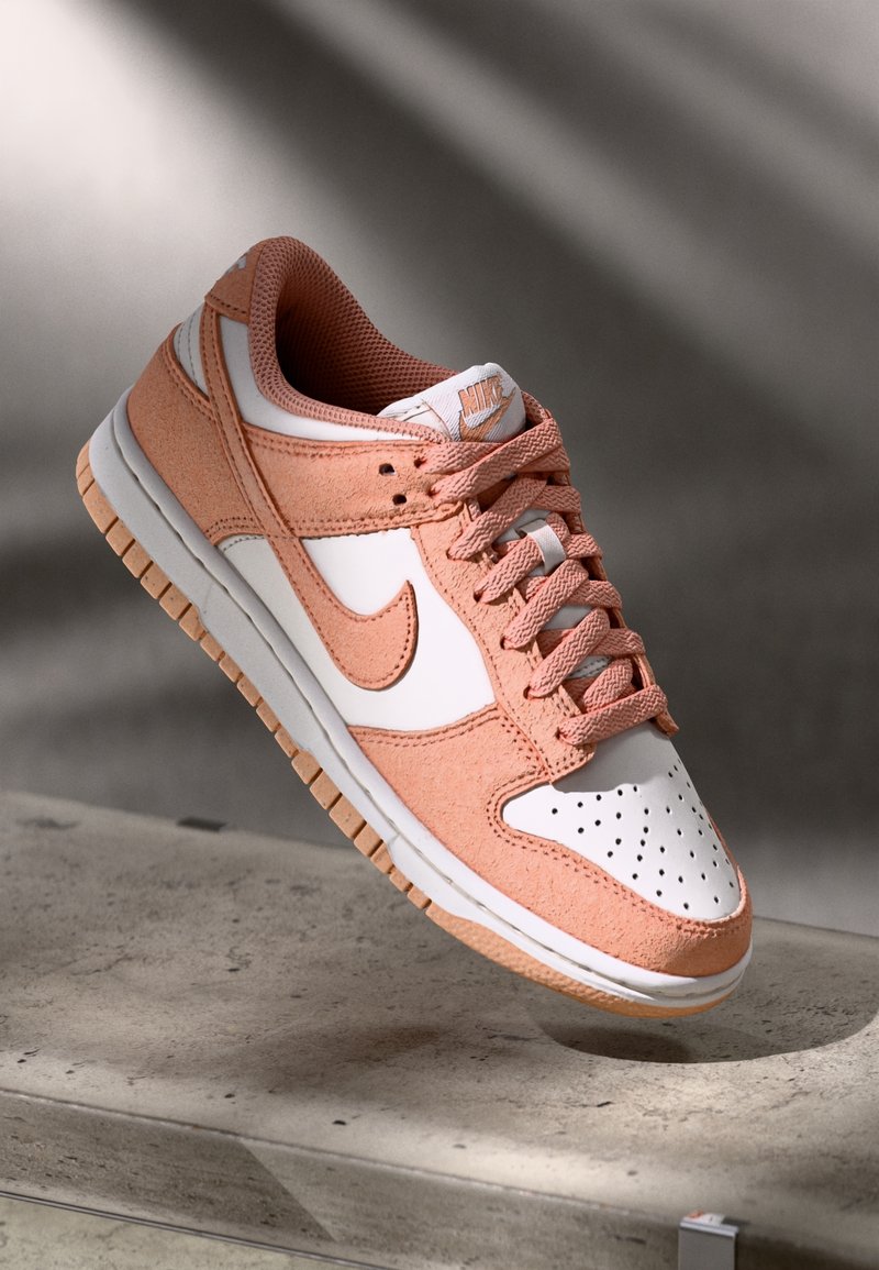 Nike Dunk Low sneakers med en vit läderbas och rosa mockaöverdrag, texturerade snören och en gummisula med mönstrat grepp.