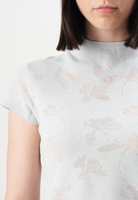 Lys blå strikket T-skjorte med korte ermer og høy hals, med et subtilt blomsterdesign i en litt mørkere nyanse. Myk, teksturert stoff.