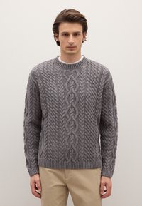 Maglione grigio lavorato a maglia con motivo a trecce, scollo rotondo e polsini e orlo a coste. Indossato sopra una camicia bianca, abbinato a pantaloni beige.
