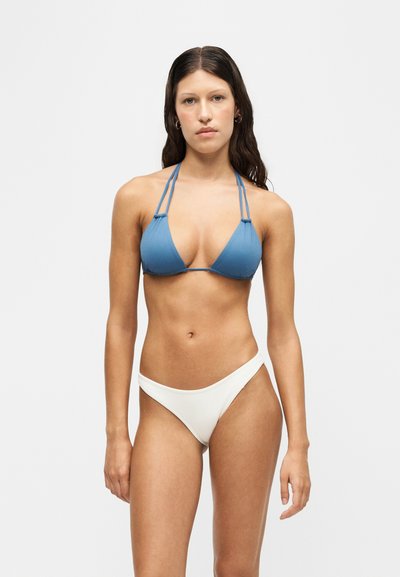 Femme aux longs cheveux foncés portant un haut de bikini bleu à col halter et un bas de bikini blanc sur un fond clair uni.