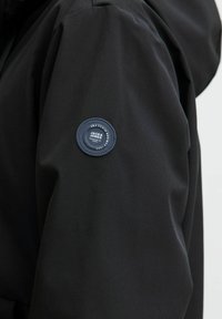 Veste extérieure noire avec une finition mate, présentant un patch en tissu rond sur la manche avec le nom de la marque et des détails. Texture douce.
