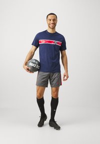 Jūrinis marškinėliai su raudona juostele ir logotipu, pilki šortai su raudonais akcentais, juodos kojinės ir juodi futbolo bateliai. Laiko pilką futbolo kamuolį.