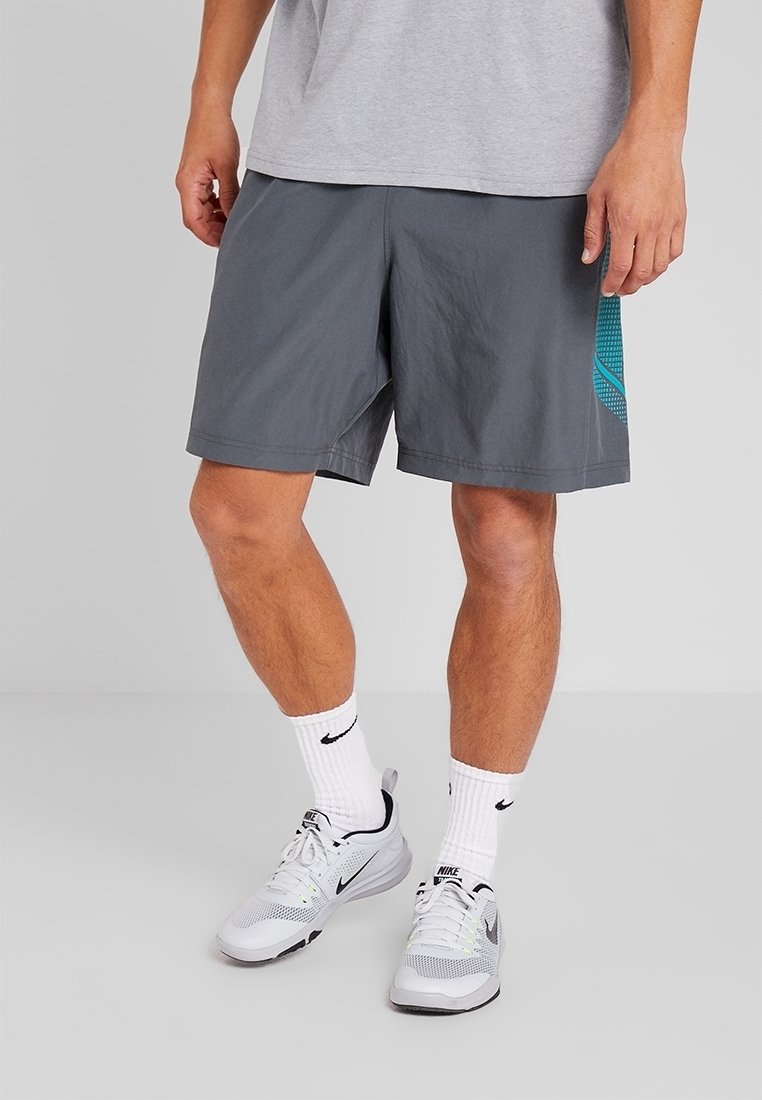 Muž v šedých sportovních šortkách, s bílými ponožkami Nike a bílými běžeckými botami Nike, stojí na světle šedé podlaze.