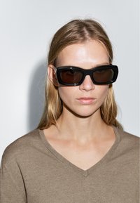 Schwarze rechteckige Sonnenbrille mit dunklen Gläsern und einem dicken Rahmen. Das Modell trägt einen beige V-Ausschnitt-Pullover vor einem schlichten Hintergrund.