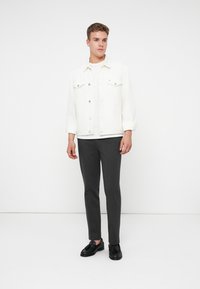 Fiatal férfi áll, fehér farmerdzsekit, fehér pólót, sötétszürke nadrágot és fekete francia cipőt visel, egy sima fehér háttér előtt.