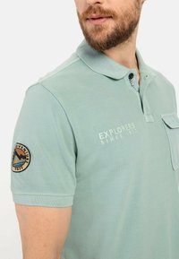 Polo shirt in lichtgroen, gemaakt van textuurstof. Voorzien van een knoopsluiting op de kraag, een borstzak en een ronde badge op de linker mouw.