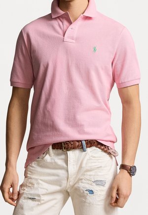 Polo shirt rosa chiaro con una finitura testurizzata e un logo ricamato verde. Abbinato a jeans bianchi strappati e una cintura intrecciata marrone.