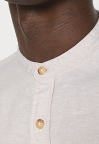 Helles beigefarbenes Leinenhemd mit rundem Kragen, versehen mit zwei beigen Knöpfen und sichtbarer Textur. Besonderer Fokus auf die Stichdetails entlang des Ausschnitts.