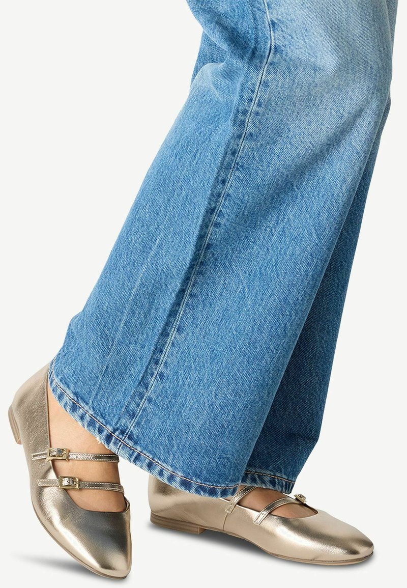 Guldmetalliska balettpumps med två ankelremmar, platt sula och spetsig tå, kombinerade med vida blå denimbyxor.