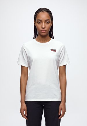 Une femme aux cheveux tressés porte un T-shirt Burton blanc et un pantalon noir sur un fond clair et uni.