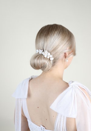 Accessorio per capelli floreale bianco con perle, posizionato in uno chignon basso. Lo sfondo è neutro e la parte superiore presenta fiocchi di tessuto bianco trasparente.