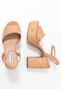 Nude suede sandály na vysokém podpatku s robustní platformou, otevřenou špičkou a nastavitelným páskem na kotníku. Význačná hladká textura a zaoblený design.