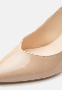 Γυναικεία μυτερή γόβα υψηλού τακουνιού από μπεζ λουστρίνι, με ομαλή επιφάνεια και λεπτομερείς ραφές κατά μήκος της άκρης του ανοίγματος.