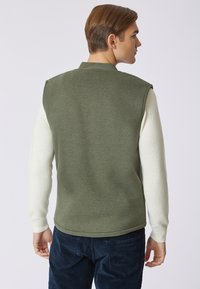 Gilet senza maniche verde oliva con una finitura texturizzata, abbinato a maniche lunghe bianche, indossato sopra pantaloni blu navy.