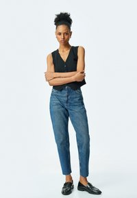 Top negro sin mangas con detalles de botones, jeans de mezclilla azul de talle alto, de longitud hasta el tobillo, de tela texturizada, combinados con mocasines negros brillantes.