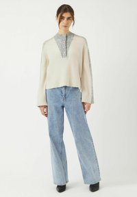 Pull en tricot beige et gris avec un col en V, des boutons décoratifs et des manches larges, associé à un jean taille haute bleu clair et des chaussures noires.