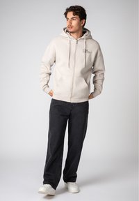 Light beige zip-up hoodie with a drawstring hood and front pockets, worn over black pants and white sneakers. Smooth texture, casual fit.