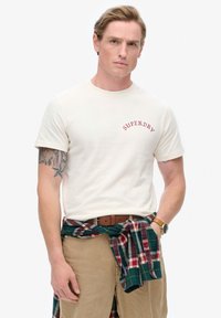 Camiseta de manga corta color crema con el logo "SUPERDRY" en rojo. Camisa de cuadros atada a la cintura en verde, rojo y azul. Pantalones beige y reloj.