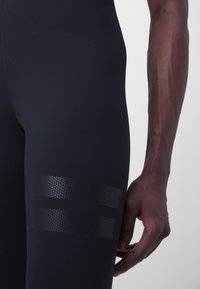 Černé sportovní legíny vyrobené z hladkého, elastického materiálu s texturovaným, perforovaným pruhovaným designem na horní části stehenního svalu.