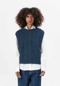 Dunkelblaues Strickweste mit Kabelmuster, gefüttert, ärmellos und mit Rundhalsausschnitt, über einem weißen Hemd getragen.
