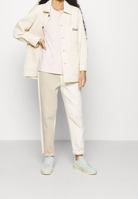 Light beige button-up jacket with front pockets over a light pink t-shirt, paired with cream and beige tapered pants and mint green sneakers.