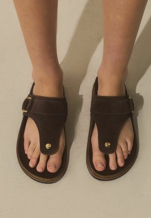 Pieds portant des sandales en suède brun foncé avec des accents dorés, debout sur une surface texturée de couleur claire.