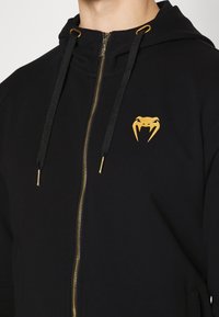 Černá mikina na zip s logem zlatého hada na hrudi, se zlatými kovovými doplňky a černými stahovacími šňůrkami. Měkká tkanina s hladkým povrchem.