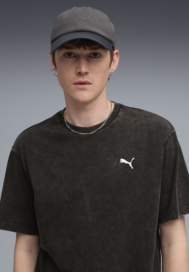 Camiseta negra de gran tamaño con textura desgastada, con un logo de Puma en blanco. Gorra gris con un diseño texturizado y visera curvada. Collar de cadena plateado.