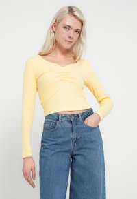 Żółta bluzka z długim rękawem o prążkowanej fakturze i dekolcie w serek, z marszczeniem na środku, zestawiona z niebieskimi jeansami z wysokim stanem.