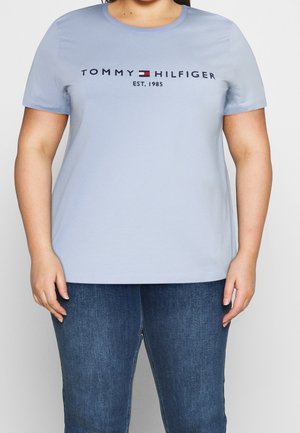 Osoba nosząca jasnoniebieską koszulkę Tommy Hilfiger z logo i napisem "EST. 1985", w połączeniu z niebieskimi dżinsami, stojąca na białym tle.