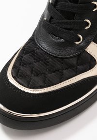 Svarta sneakers med en quiltad textur på ovandel, läderdetaljer, guldfärgad metallfinish och runda svarta snören för fastsättning.